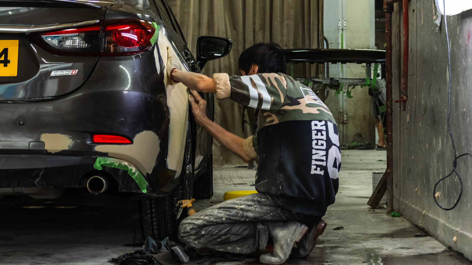 A person working beside the rear panel of a gray car in a repair workshop.