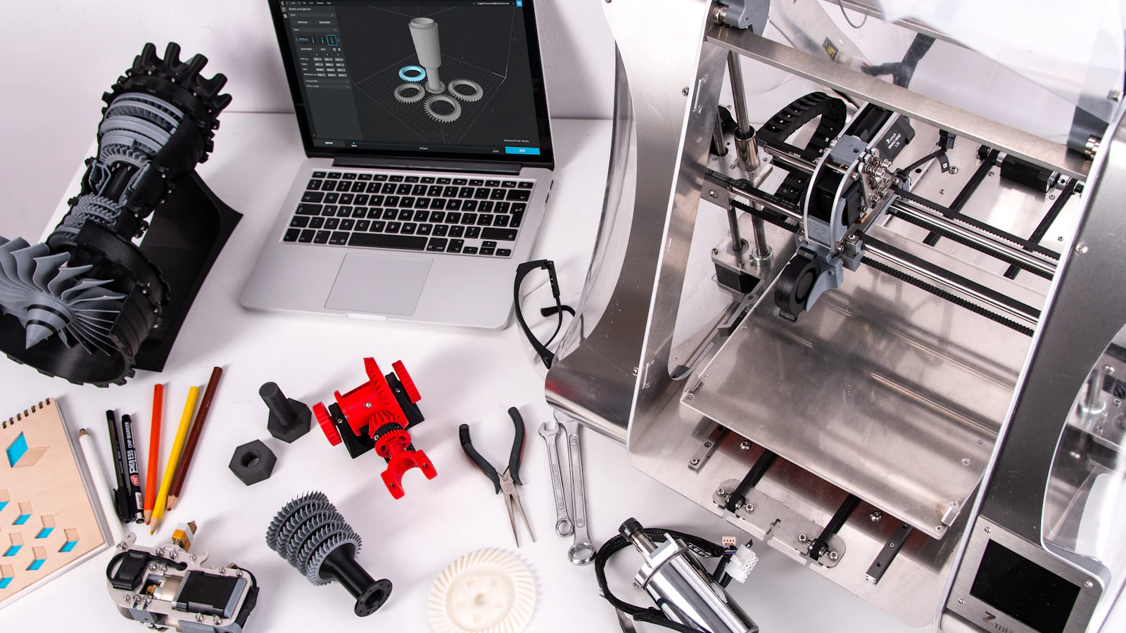 A laptop, tools and machine parts on a worktable.