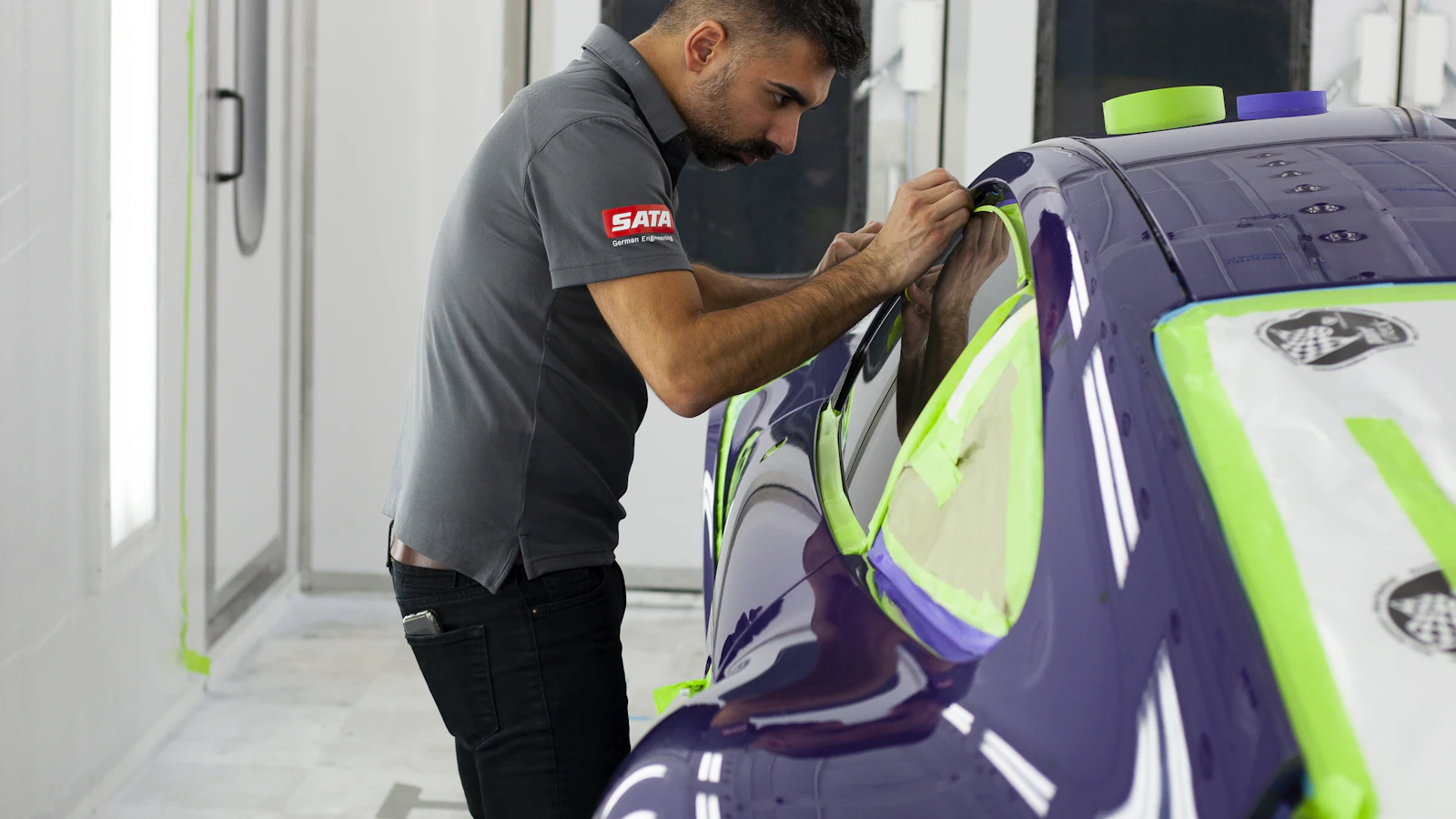 A person applying masking tape to a purple vehicle panel in a workshop.