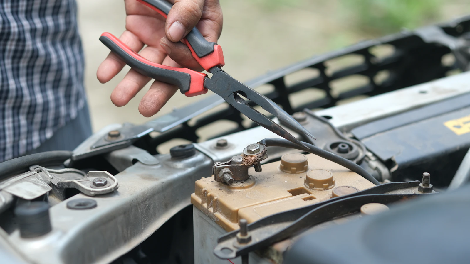 A hand holding red-handled pliers above vehicle engine components.