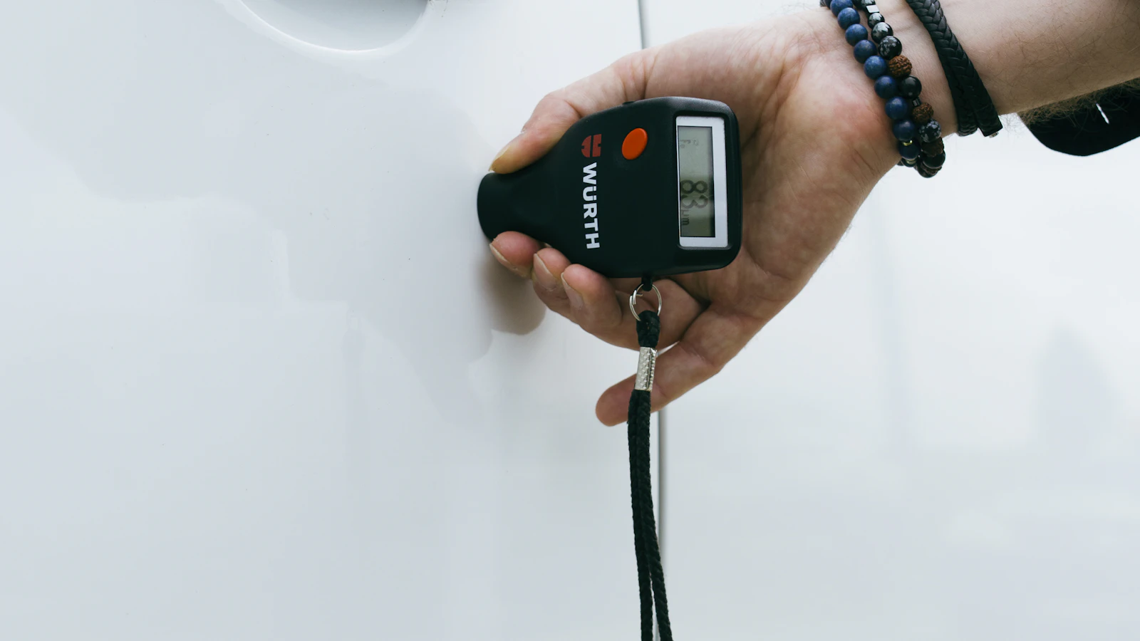 A hand holding an inspection meter beside a white vehicle panel.