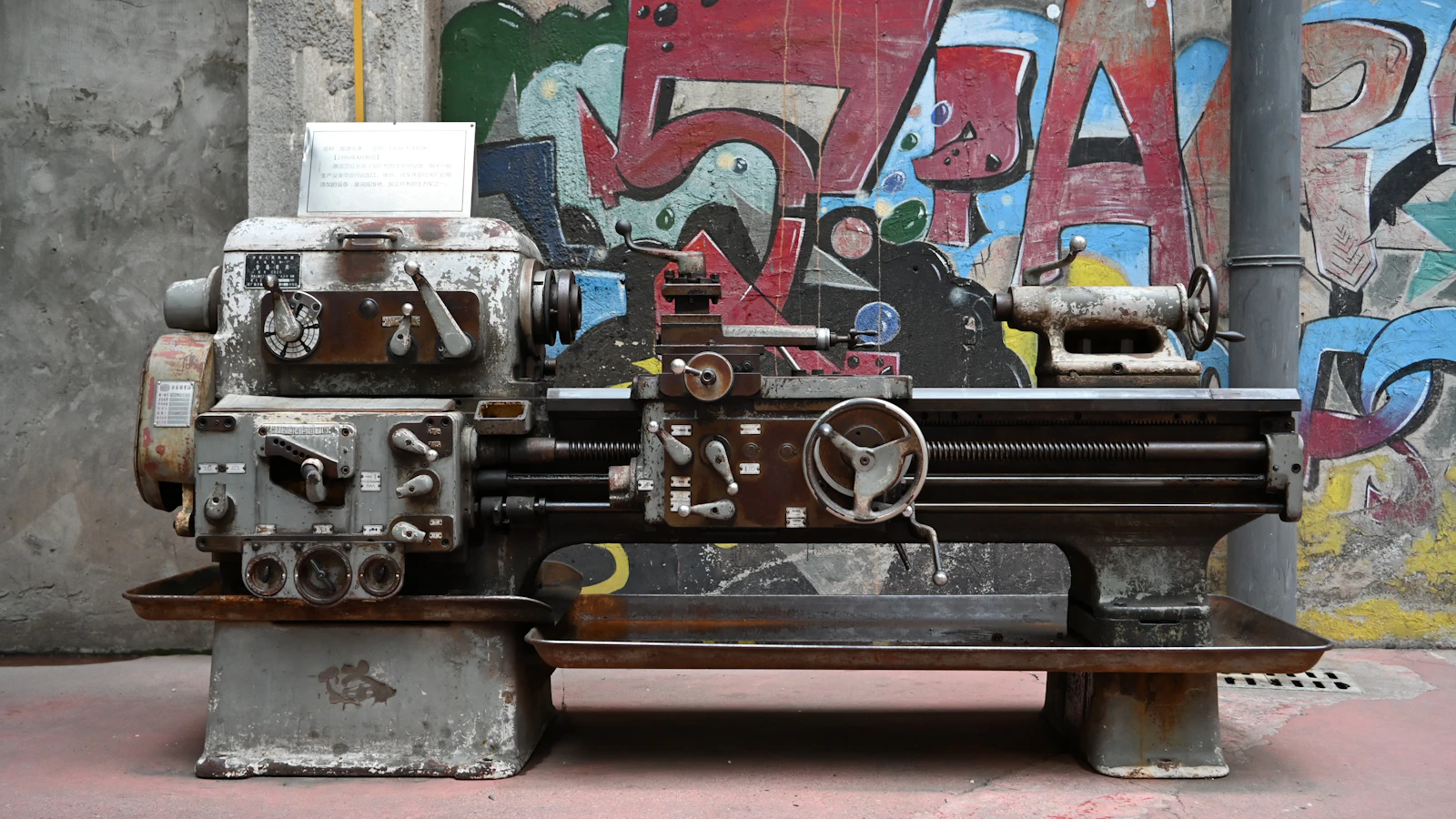 A metal lathe standing in front of a graffiti-covered workshop wall.