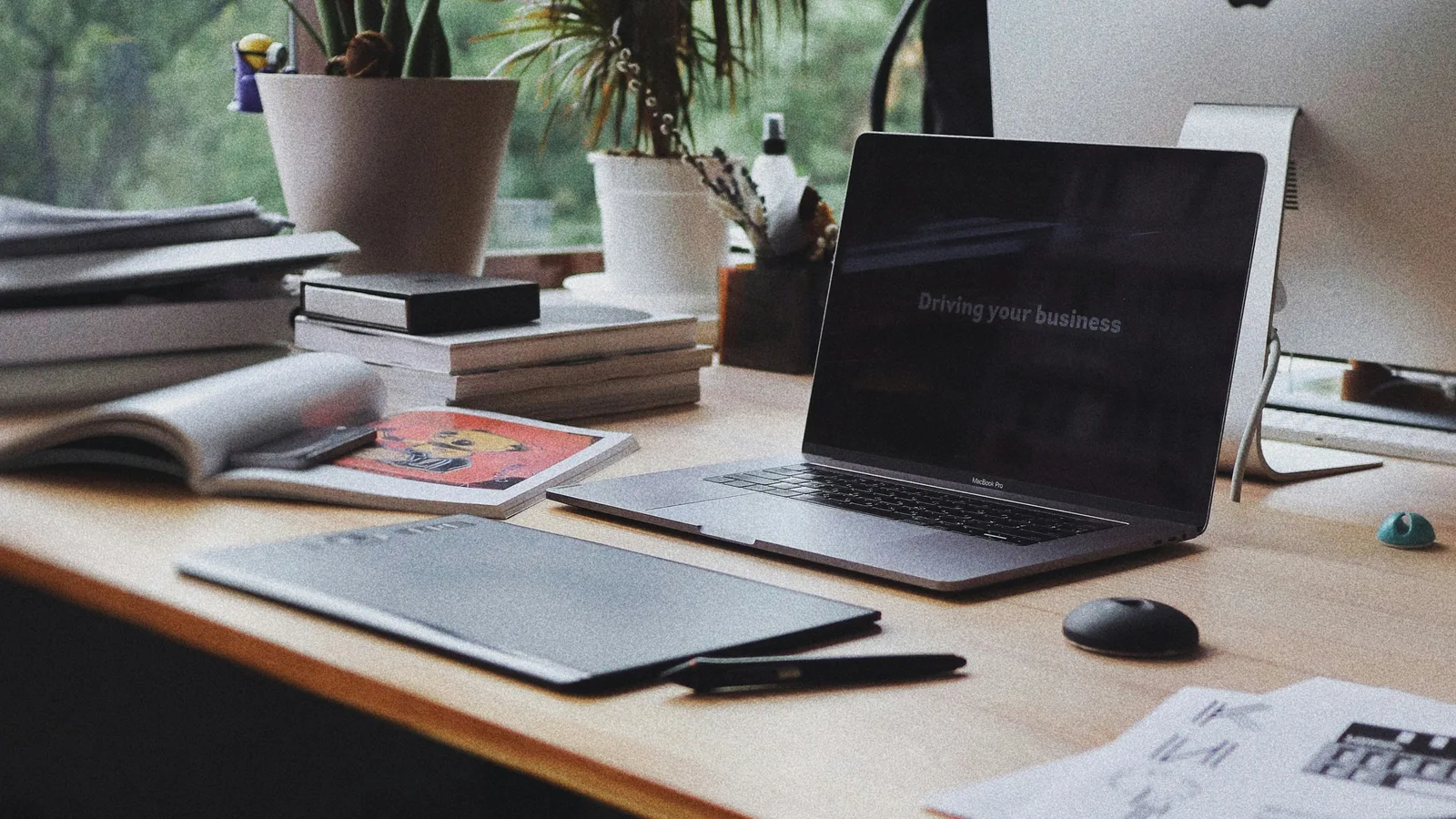 Laptop, books and an open magazine on a desk by a window.
