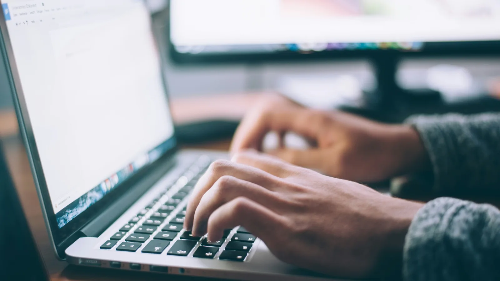 Hands typing on a laptop beside a desktop monitor.
