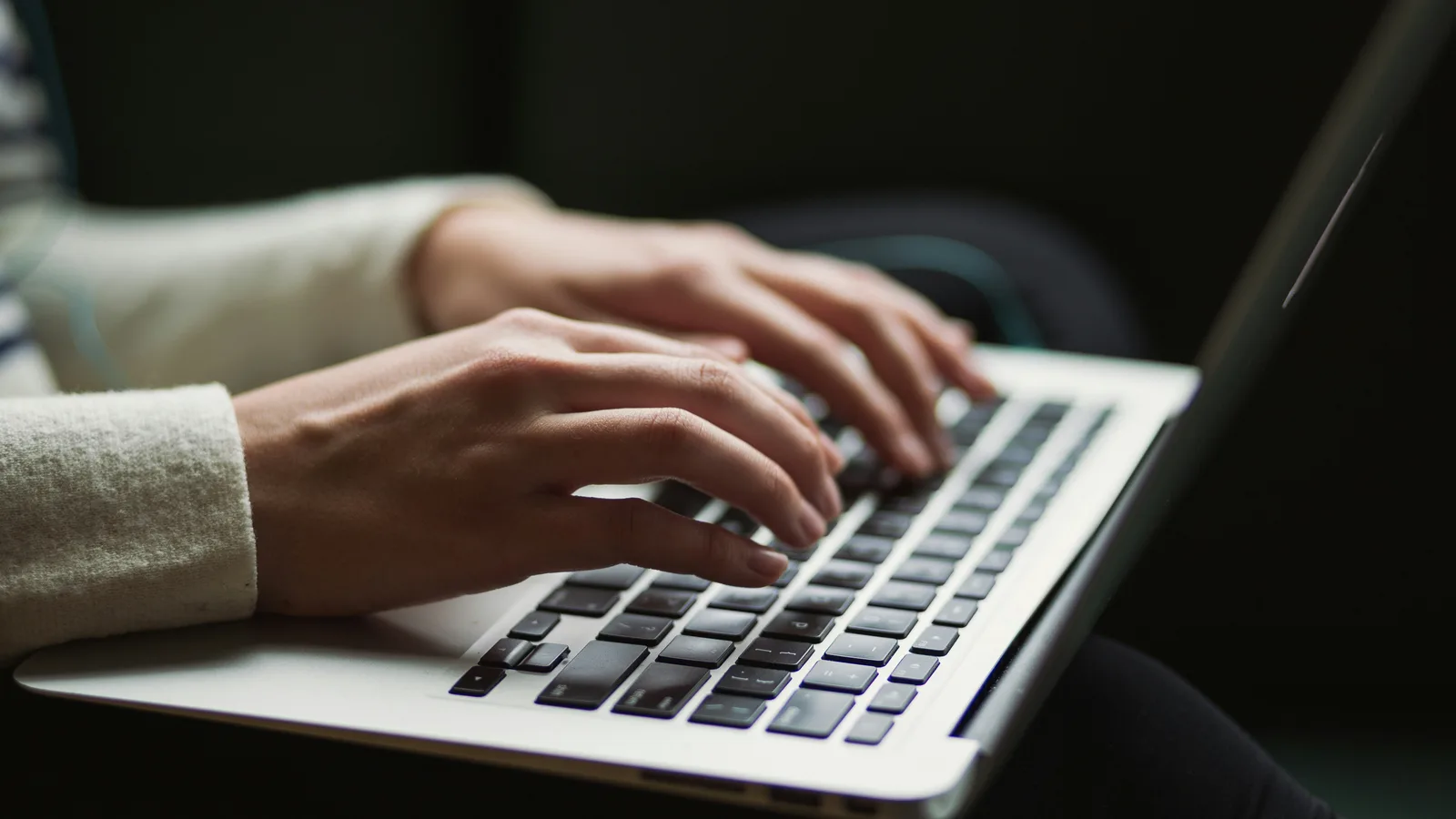 Close-up of hands typing on a laptop in a dim room.