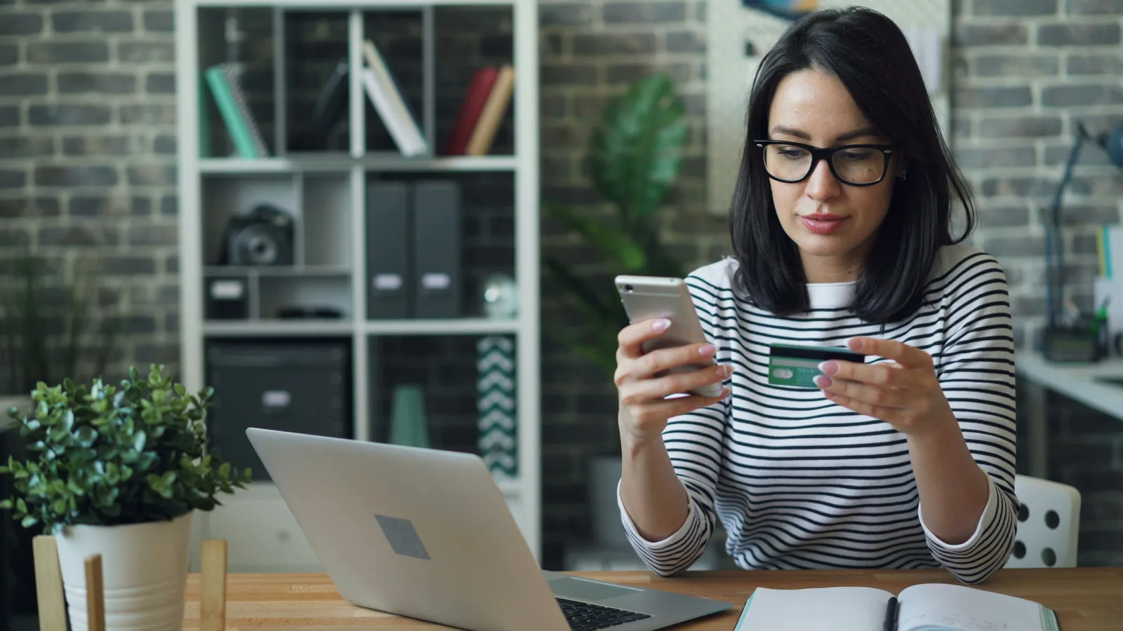 Woman checking a payment card while using a smartphone beside a laptop.