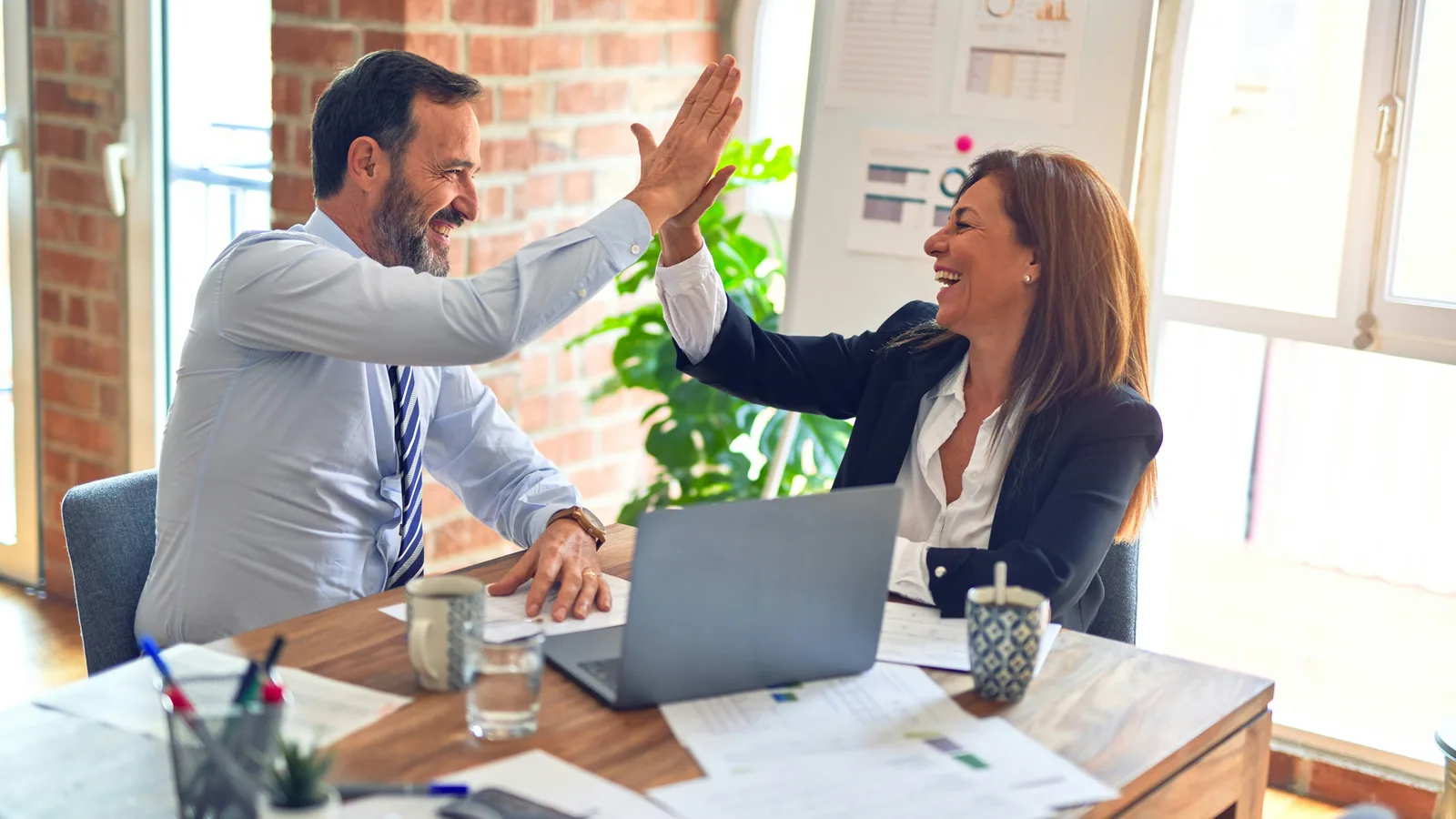 Two colleagues exchanging a high five across a desk with a laptop.