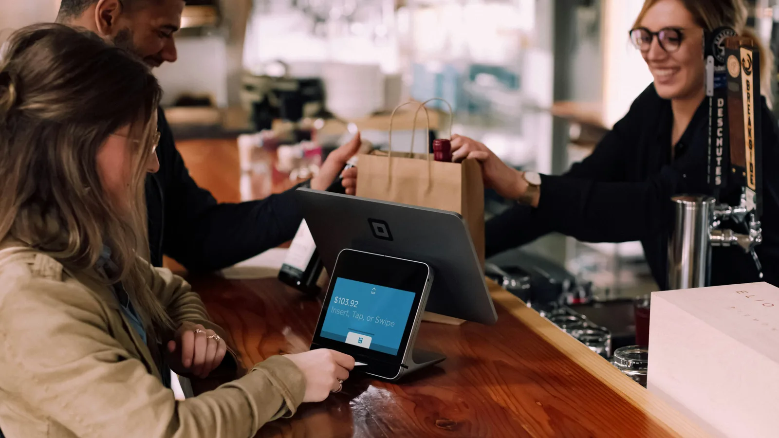 Customer using a checkout screen while a staff member hands over a shopping bag.