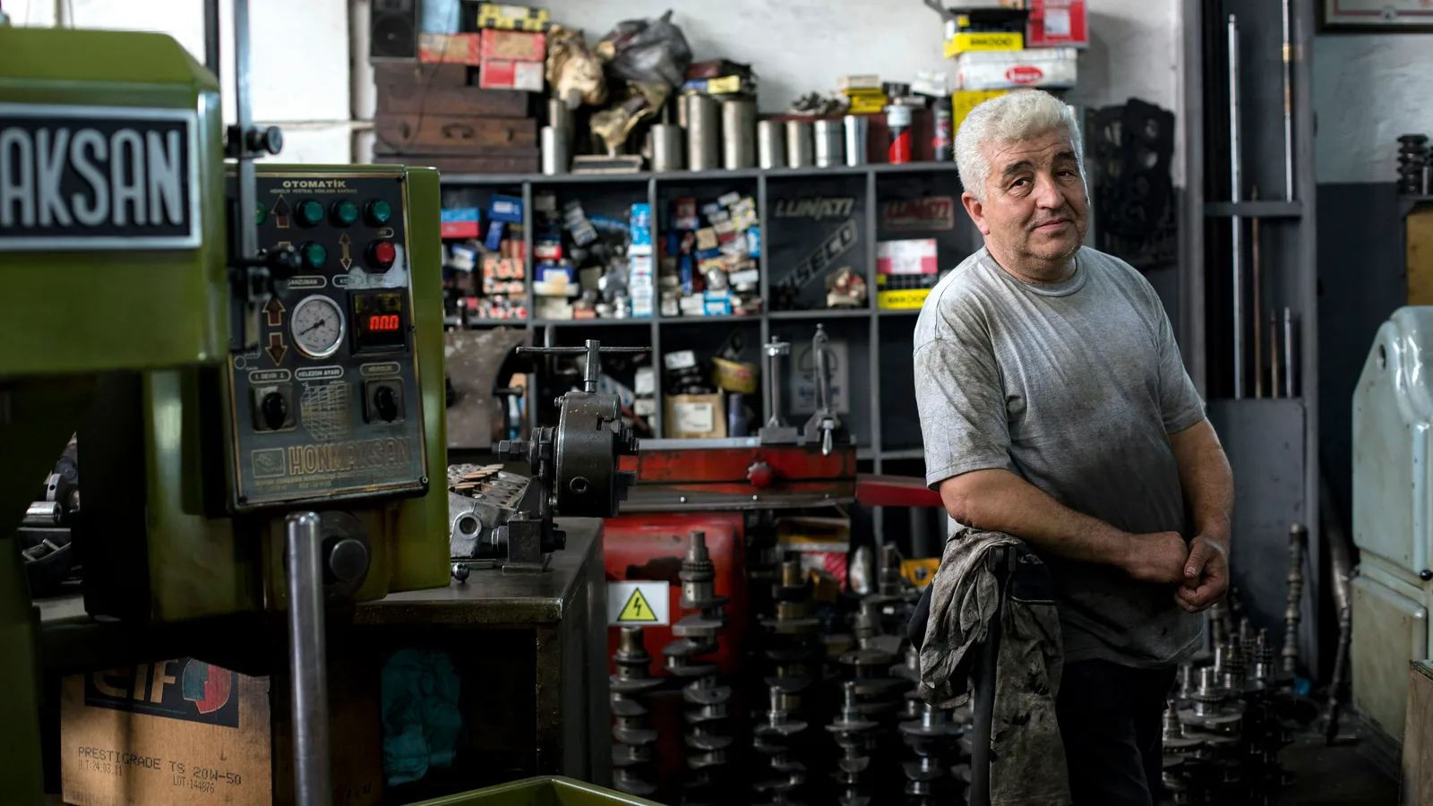 Man standing beside machinery and shelves of parts in a workshop.