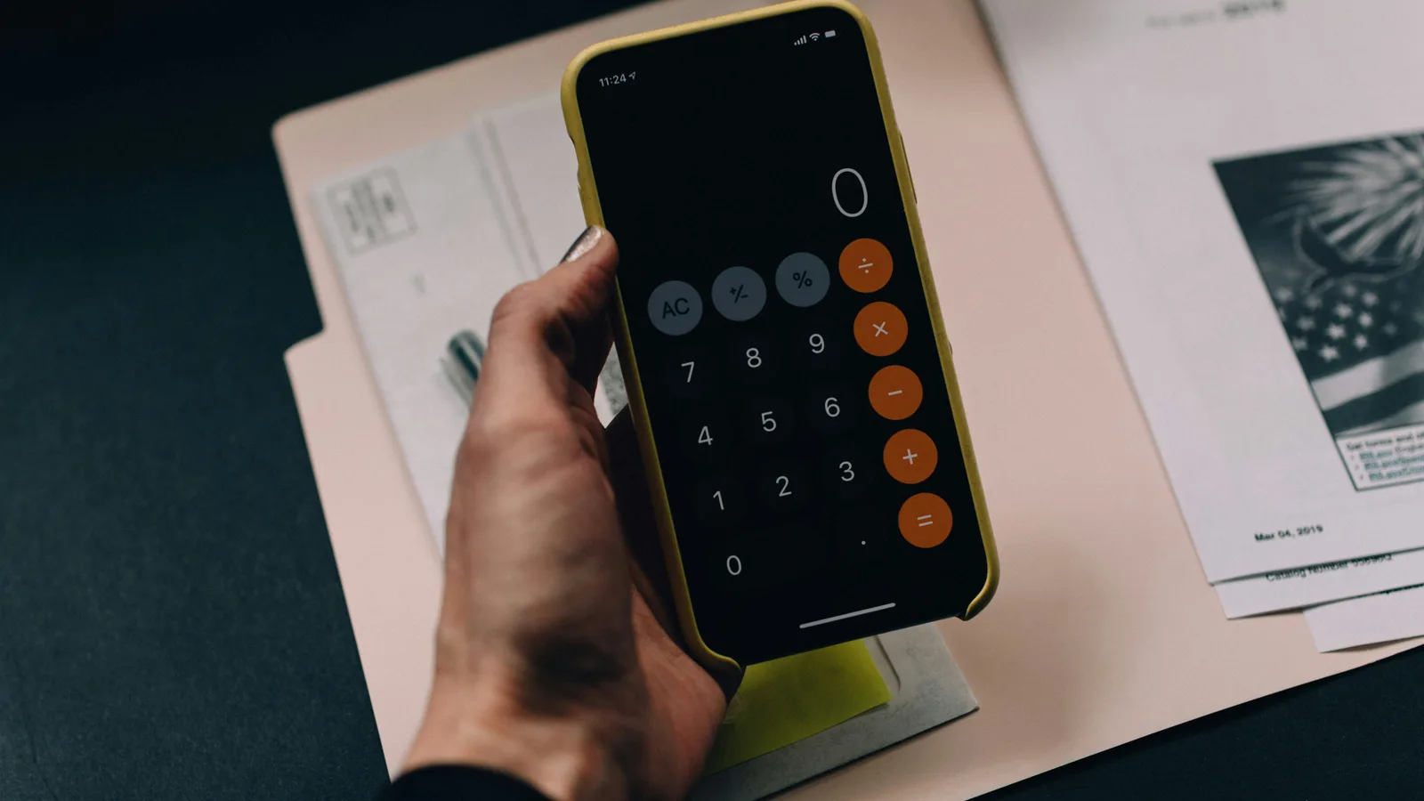 A hand holding a smartphone displaying a calculator app over a paper folder.