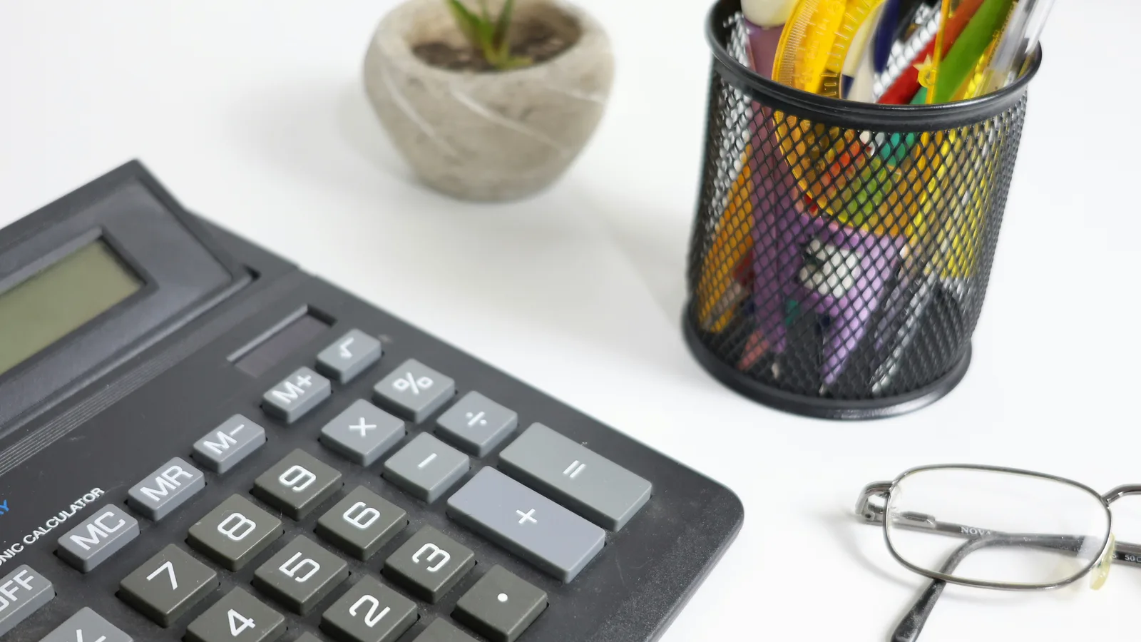 Calculator, glasses, and pens on a white desk.