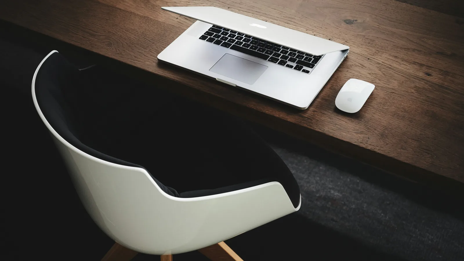 Laptop and mouse on a wooden desk beside a white chair.
