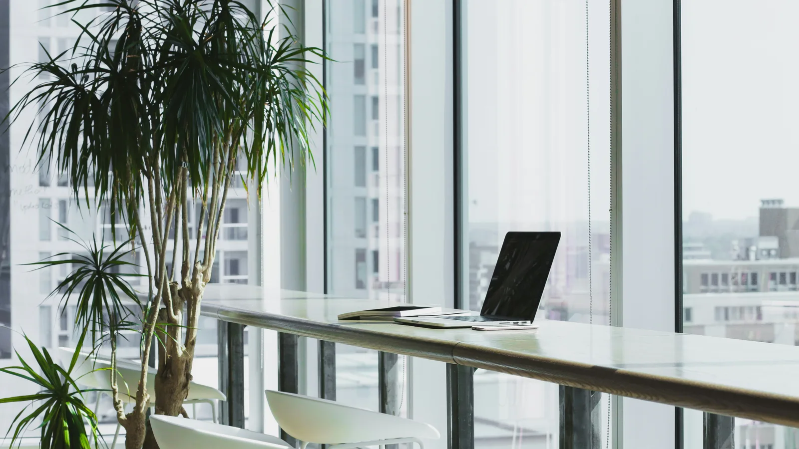 Laptop on a long desk beside tall office windows.