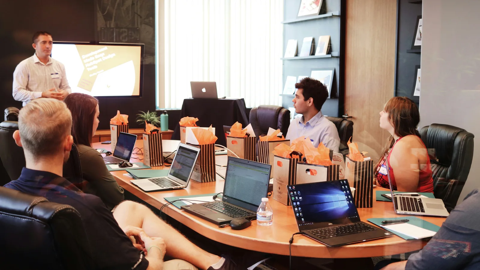 Presenter speaking to colleagues seated around a table with laptops.