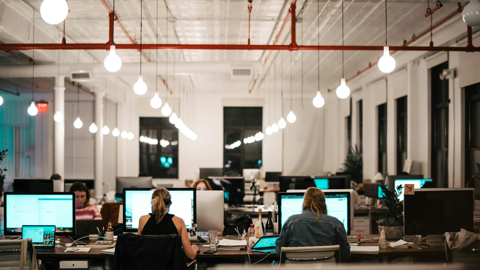 People working at computers beneath exposed ceiling beams and hanging lights.