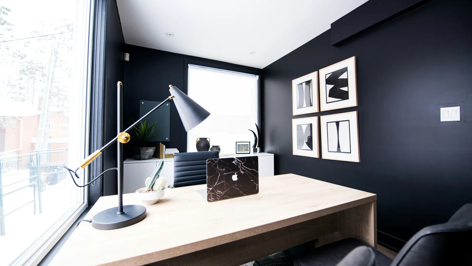 Desk with a laptop, lamp and framed art beside a window.