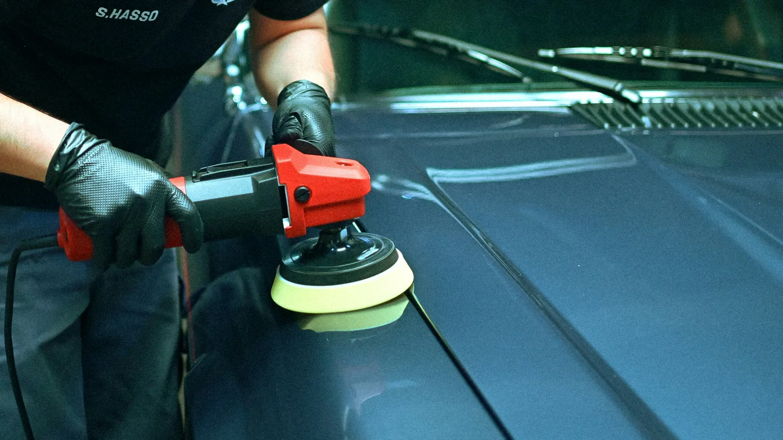 Gloved hand polishing the hood of a blue car with a powered buffer.